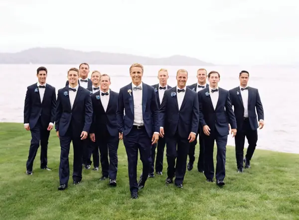 Groomsmen in navy suits with bow ties walk on grass by a lake. Can you wear a normal suit to a black tie event? This image shows a stylish alternative.