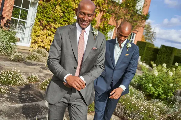 Two men in suits walk outdoors. The question "do you have to wear a suit to a wedding?" is answered by their stylish attire.